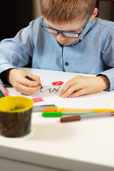 Chłopiec w niebieskiej koszuli i okularach siedzi przy białym biurku i rysuje kredkami na białej kartce papieru. Na pierwszym planie kolorowe flamastry. - obrazy, fototapety, plakaty