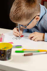 Chłopiec w niebieskiej koszuli i okularach siedzi przy białym biurku i w skupieniu rysuje cienkopisami na białej kartce papieru. Na pierwszym planie kolorowe flamastry. - obrazy, fototapety, plakaty