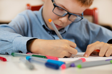 Chłopiec w niebieskiej koszuli i okularach siedzi przy białym biurku i w skupieniu rysuje cienkopisami na białej kartce papieru. Na pierwszym planie kolorowe flamastry. - obrazy, fototapety, plakaty