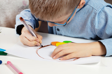 Chłopiec w niebieskiej koszuli i okularach siedzi przy biurku i w skupieniu wykonuje pracę plastyczną z kolorowego papieru. Chłopiec maluje brązowym flamastrem. - obrazy, fototapety, plakaty
