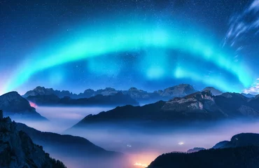 Selbstklebende Fototapeten Aurora borealis above mountains in fog at night. Northern lights. Sky with stars with polar lights and high rocks. Beautiful landscape with aurora, city lights in low clouds, mountain ridge. Space © den-belitsky