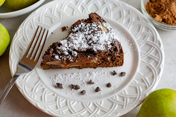Close up top side view of Chocolate Pear Cake.  Chocolate chips and fork on plate.  Sprinkled with powdered sugar.  Pears and cocoa powder in background.