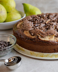 Close up view of left side of Chocolate Pear Cake with measuring spoons and chocolate chips in foreground.  Bowl of pears in background.