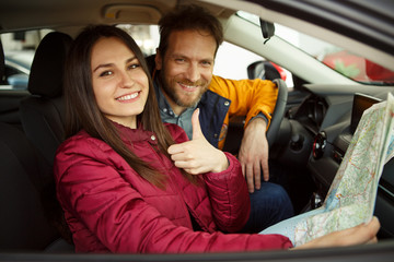 Fototapeta na wymiar Pretty couple sitting in automobile cabin, smiling and looking at camera. Woman and man buying new car and planning trip. Beautiful woman holding map and showing thumb up.