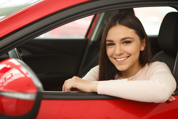 Fototapeta na wymiar Pretty girl with brilliant smile looking at camera from window of red car. Happy young woman sitting in vehicle and enjoying comfortable automobile cabin. Purchase of new auto in car center.