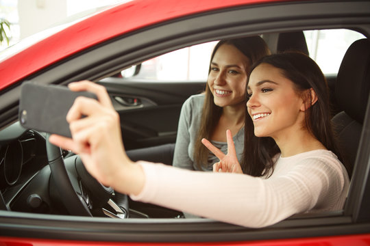 Two Pretty Girls Making Selfie In Automobile Cabin. Girls Sitting Inside, Smiling, One Girl Holding Phone. Female Customers Happy Because Of Purchasing New Auto In Car Center.