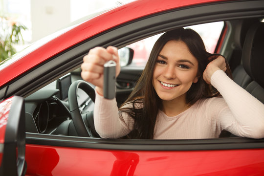 Side view of beautiful girl smiling and looking at camera. Gorgeous young woman holding keys of her new red auto. Happy female client of car dealership buying automobile.