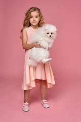 Little girl with a blond curly hair, in a pink dress plays with her dog