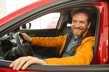 Red haired man enjoying comfortable seats of new red car. Male want buy new automobile in modern car dealership, sitting in vehicle, smiling and looking at camera. Concept of transportation.