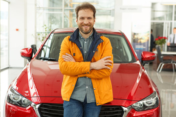 Positivity and handsome man customer posing with crossed hands in showroom of car center where he buy his new chic automobile. Male owner leaning of new red car, looking, smiling at camera.