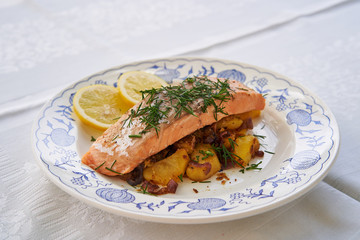 Salmon fillet or steak served with roasted potatoes and onion, slices of fresh lemon and chopped fresh dill on the rustic plate. Fresh, healthy, light, diet fish dish for lunch or diner.