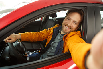 Showroom of car center. Happy bearded man sitting inside automobile and making selfie. Male customer holding hand on steering wheel and testing car before purchasing.