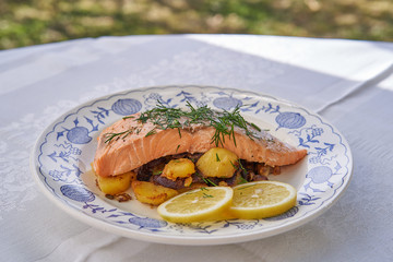 Close up picture baked salmon fillet with roasted potatoes, onion and slices of lemon and chopped fresh dill herbs served on rustic plate outdoor in garden restaurant during sunny spring day
