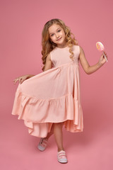 Little girl with a blond curly hair, in a pink dress is posing with a candy