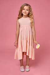 Little girl with a blond curly hair, in a pink dress is posing with a candy