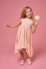 Little girl with a blond curly hair, in a pink dress is posing with a candy