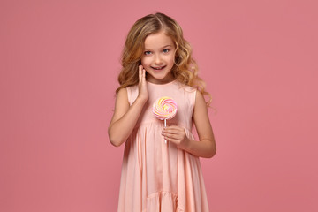 Little girl with a blond curly hair, in a pink dress is posing with a candy