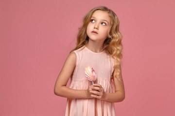 Little girl with a blond curly hair, in a pink dress is posing with a candy