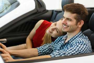 Side view of happy couple testing future car in auto salon before purchase. Smiling man on seat of driver keeping hands on wheel and looking forward and his girlfriend on passenger seat hugging him.