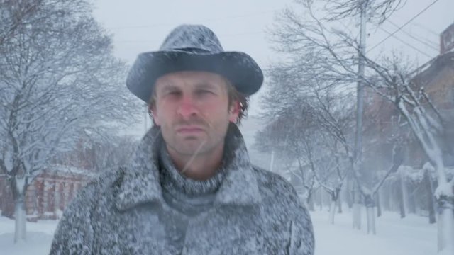 Serious man with coat and hat walks on the winter street at snowfall time close-up