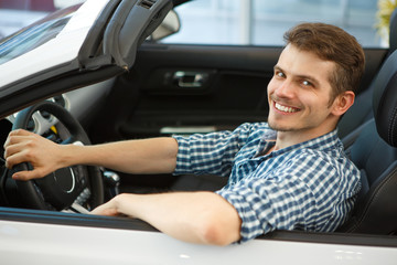 Side view of smiling handsome customer testing automobile in auto salon. Glad man wearing checkered shirt sitting in white cabriolet, keeping one hand on wheel and other on door and looking at camera.
