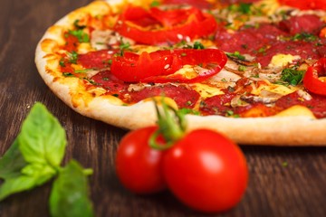 Close up Italian pizza with melted cheese, sausage, ham, salami, mushrooms, pepper and fresh herbs on a brown table decorated by mushrooms, red sweet pepper and cherry tomatoes