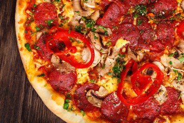 Close up top view Italian pizza with melted cheese, sausage, ham, salami, mushrooms, pepper and fresh herbs on a brown table decorated by mushrooms, red sweet pepper and cherry tomatoes