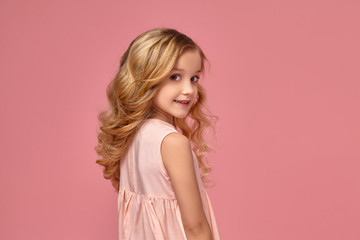 Little girl with a blond curly hair, in a pink dress is posing for the camera