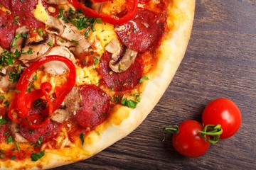 Close up top view Italian pizza with melted cheese, sausage, ham, salami, mushrooms, pepper and fresh herbs on a brown table decorated by mushrooms, red sweet pepper and cherry tomatoes