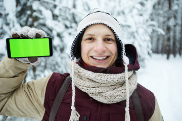 Obraz premium Portrait of active young man showing smartphone with green screen during hike in winter forest