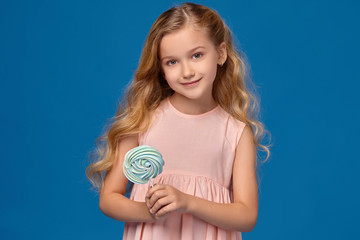 Fashionable little girl in a pink dress, with a candy in her hands, standing on a blue background.