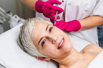 Mature woman is getting a rejuvenating facial injections. She is lying calmly at clinic. The expert...
