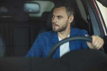 Man sitting in car, holding one hand at wheel and looking at side.