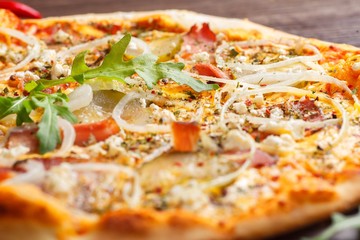 Close up Italian pizza with melted cheese, mozzarella, ham, pickled cucumbers, onion and fresh arugula leaves on a brown table decorated by mushrooms, red sweet pepper and cherry tomatoes