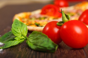 Close up top view Italian pizza with melted mozzarella cheese, red cherry tomatoes, ham and fresh green herbs on a brown table decorated by mushrooms, red sweet pepper and cherry tomatoes
