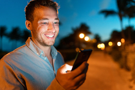 Man smiling looking at phone outside on city street at night texing online using smartphone. Young male face lit by screen light using mobile cellphone outdoors in the dark in summer.