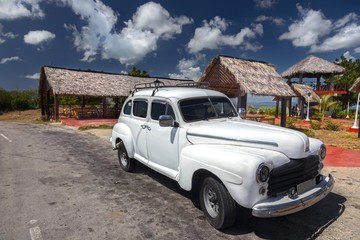 Vintage Old White Cuban Car against the background of Open Houses and Blue Sky dotted with puffy Clouds in Guantanamo Province, Cuba
