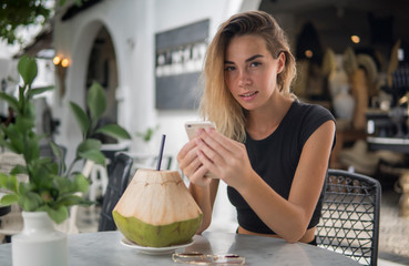 Portrait of well-graced blonde sitting in outdoor exotic cafe, coconut cocktail and fashionable sunglasses are on the table, modern white telephone in her hands, copy space