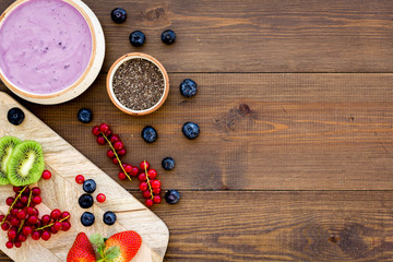 Cook trendy breakfast. Acai smoothie bowl near cutting board with fresh fruits, berries, chia seeds on dark wooden background top view copy space