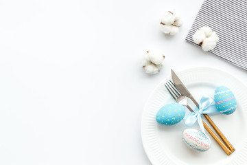 Fototapeta premium Easter table decoration in blue colors. Plate, cutlery, painted eggs and dry cotton flowers, tablecloth on white background top view space for text