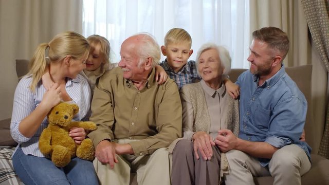 Lockdown of family members of different generations sitting together on couch in living room, talking to each other, then looking at camera and smiling