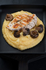 Polenta with fried chicken breast fillet and mushrooms in a cast-iron serving pan, closeup