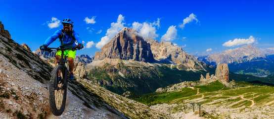 Fototapeta na wymiar Tourist cycling in Cortina d'Ampezzo, stunning Cinque Torri and Tofana in background. Man riding MTB enduro flow trail. South Tyrol province of Italy, Dolomites.
