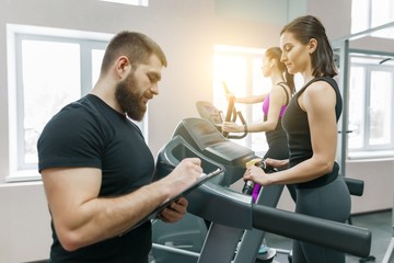 Obraz premium Young smiling fitness women with personal trainer an adult athletic man on treadmill in the gym. Sport, teamwork, training, healthy lifestyle concept