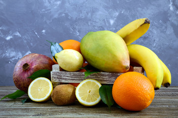 A pile of exotic fruits (bananas, oranges, kiwi, pomegranate, mango, guava, lemon) in a wooden box on the table.