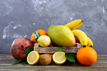 A pile of exotic fruits (bananas, oranges, kiwi, pomegranate, mango, guava, lemon) in a wooden box on the table.