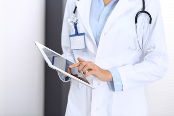 Doctor woman using tablet in clinic office. Unknown female physician at work while standing straight at hospital, close-up. Medicine and healthcare concept