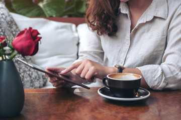 Closeup image of a woman's hands holding and pointing at a black tablet pc with coffee cup on wooden table in cafe