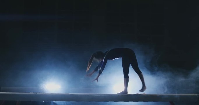 The girl performs a trick on a log in backlight and slow motion in sports gymnastic clothing. Smoke and blue. Jump and spin on the balance beam