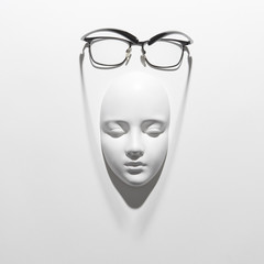 Plaster face mask with elegant glasses above it on a white background with soft long shadows, copy space. Flat lay.